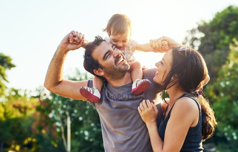 Smiling father carrying baby on shoulders, with mother smiling next to them, outdoors in bright sunlight.